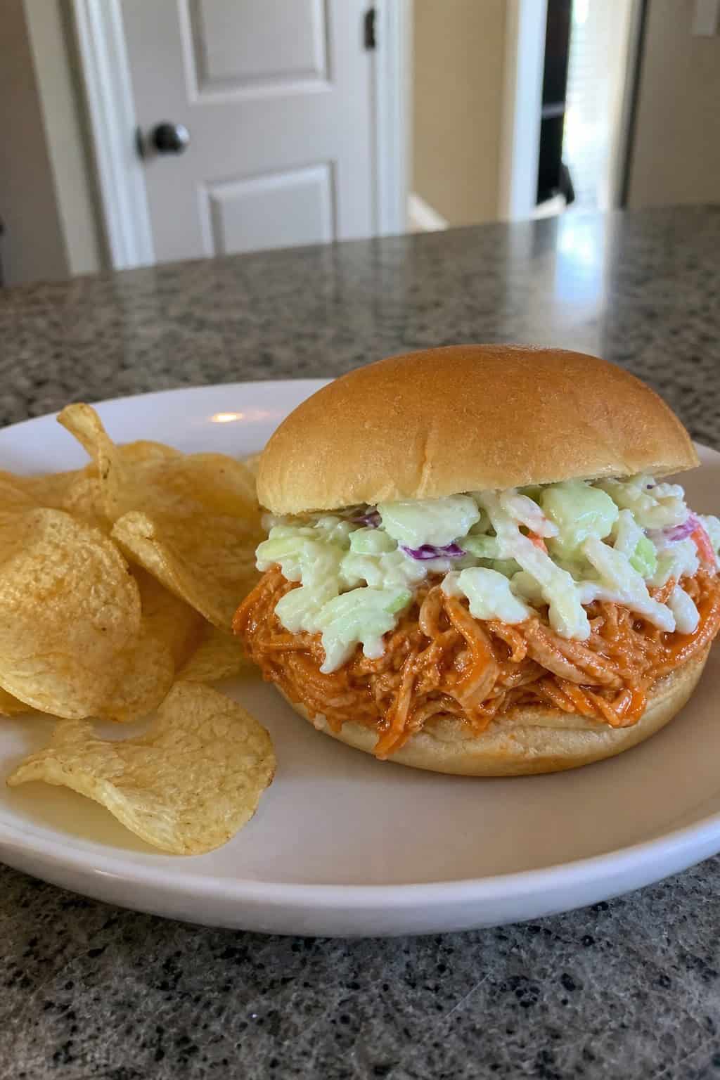 A wooden table with toasted buns piled high with saucy buffalo shredded chicken, lettuce, and a side of celery sticks and blue cheese dipping sauce.