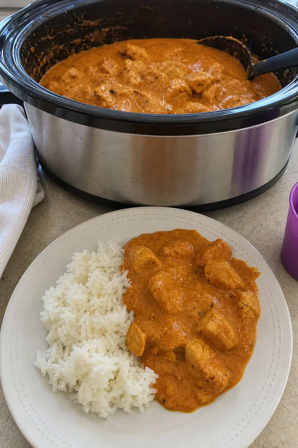A slow-cooker filled with creamy butter chicken on a wooden table surrounded by fresh ingredients and natural light.