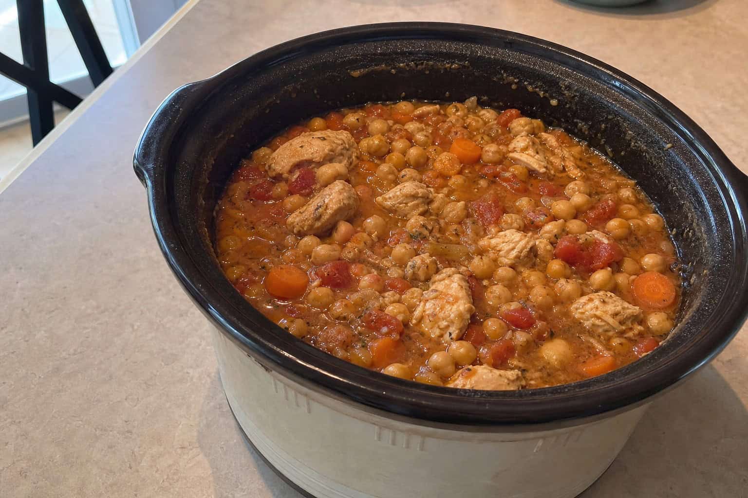 A slow cooker filled with chicken and chickpea stew on a wooden countertop surrounded by fresh ingredients.