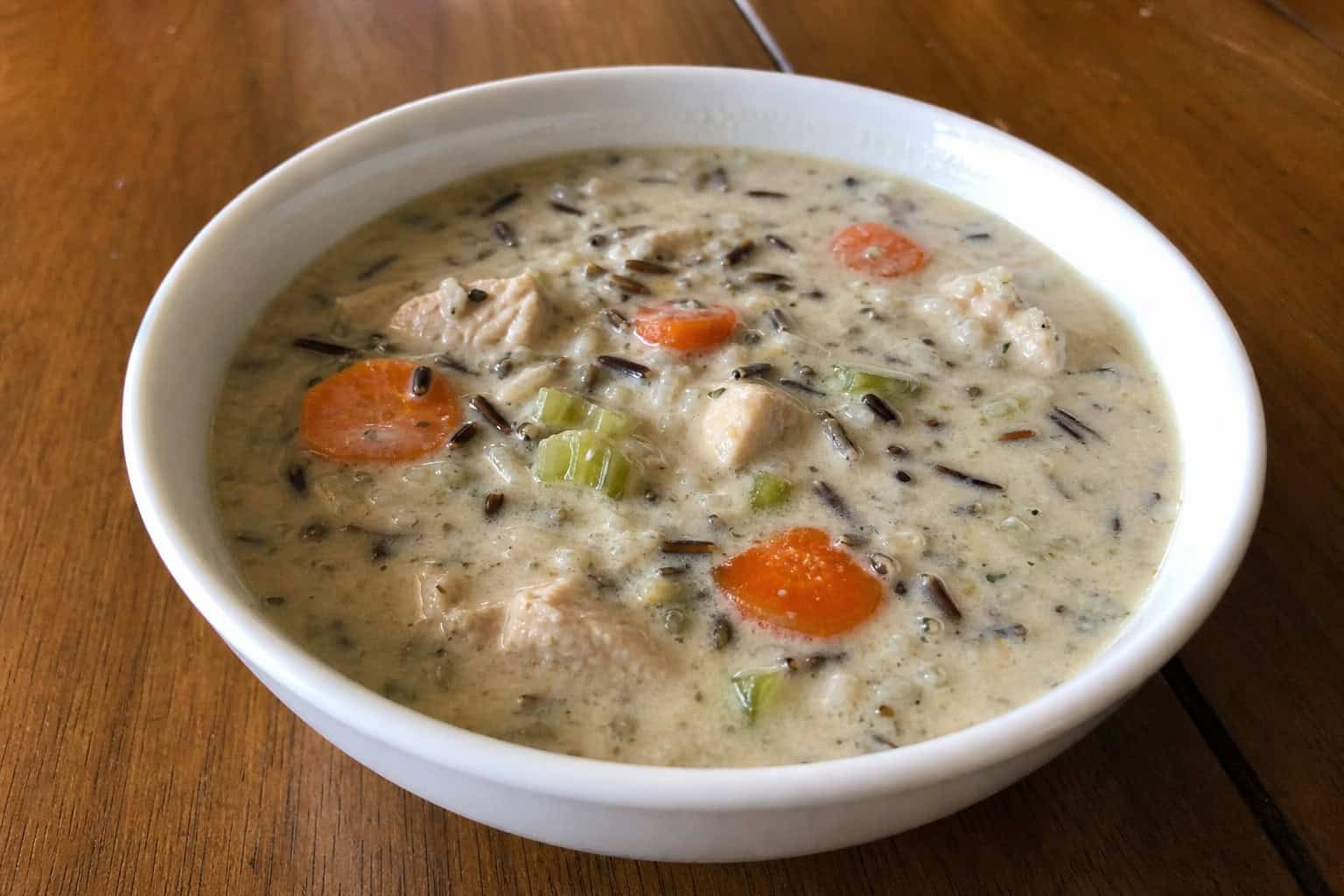 A bowl of slow cooker chicken and wild rice soup with vegetables on a wooden table, surrounded by fresh ingredients.