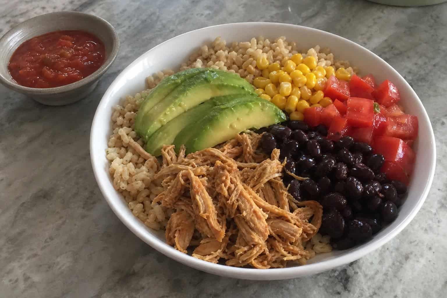 A bowl of slow cooker chicken burrito with rice, black beans, avocado, tomatoes, corn, and lime on a wooden table.