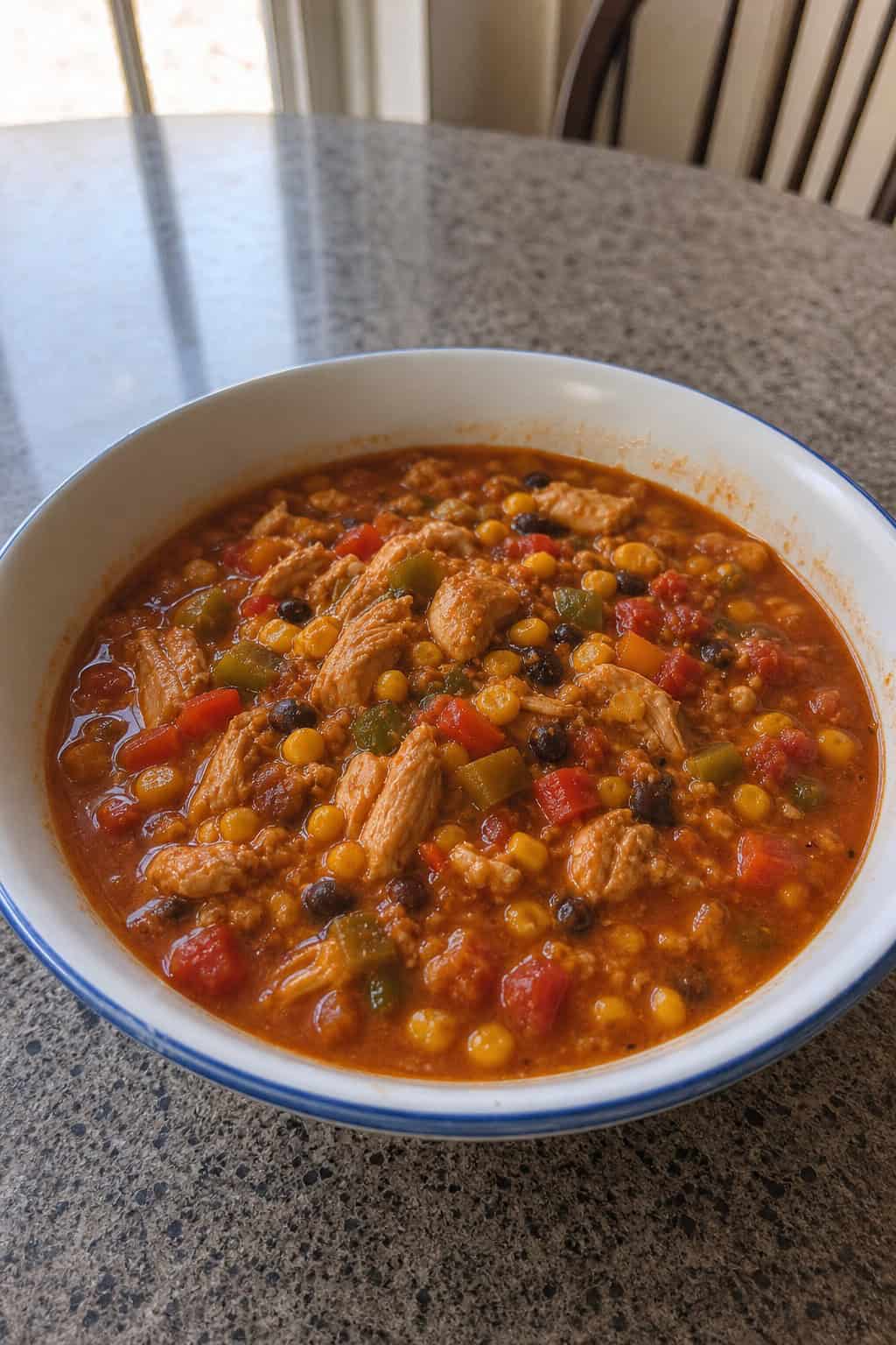 A slow-cooker filled with chicken chili containing corn and colorful bell peppers on a wooden kitchen countertop with fresh vegetables nearby.