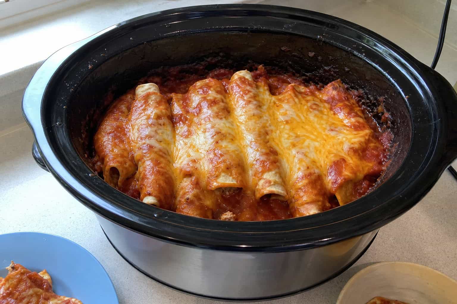 Slow-cooker filled with chicken enchiladas surrounded by fresh ingredients on a kitchen counter.