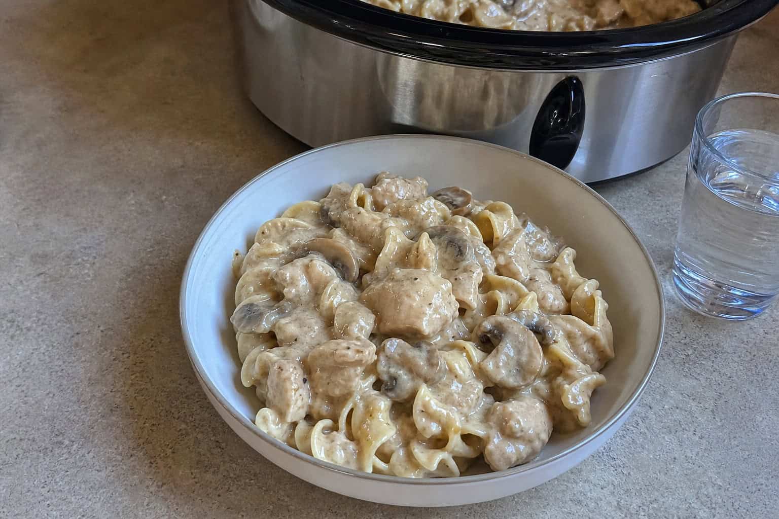 A bowl of creamy chicken stroganoff with mushrooms on a wooden table next to a slow cooker and fresh ingredients.