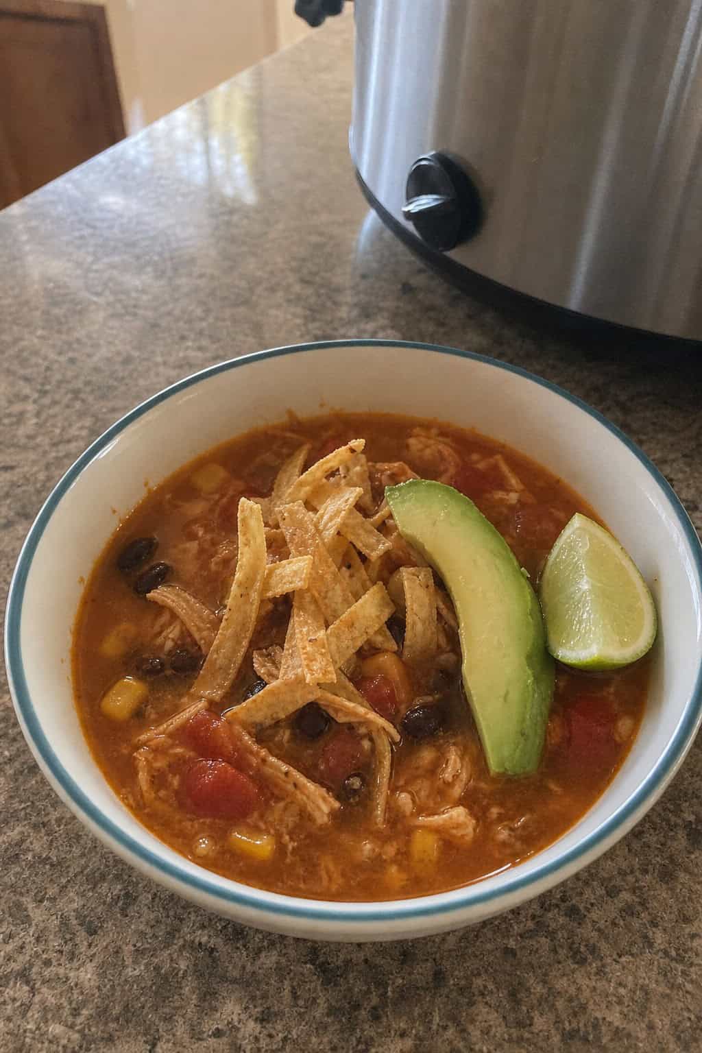 A bowl of chicken tortilla soup with tortilla strips, avocado, and cilantro on a wooden table surrounded by fresh ingredients and a slow cooker.