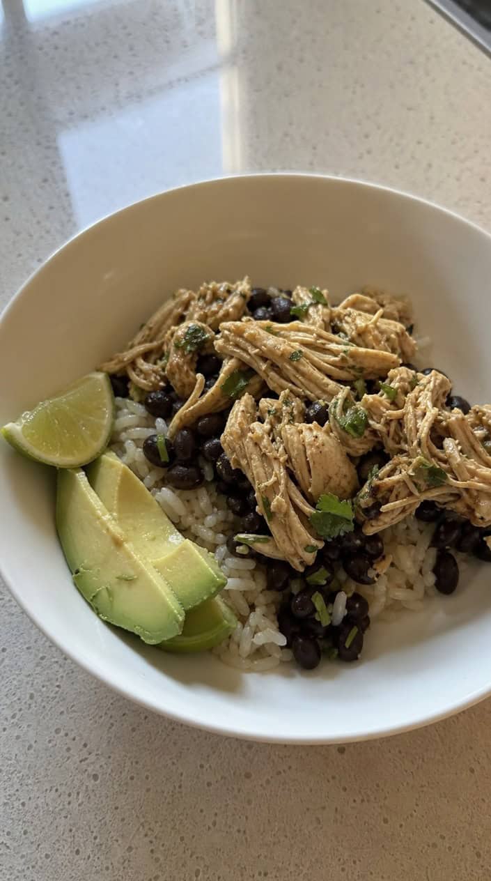 A deep bowl of cilantro-lime shredded chicken over white rice and black beans, topped with lime wedges and a scatter of fresh cilantro on a warm wooden table.