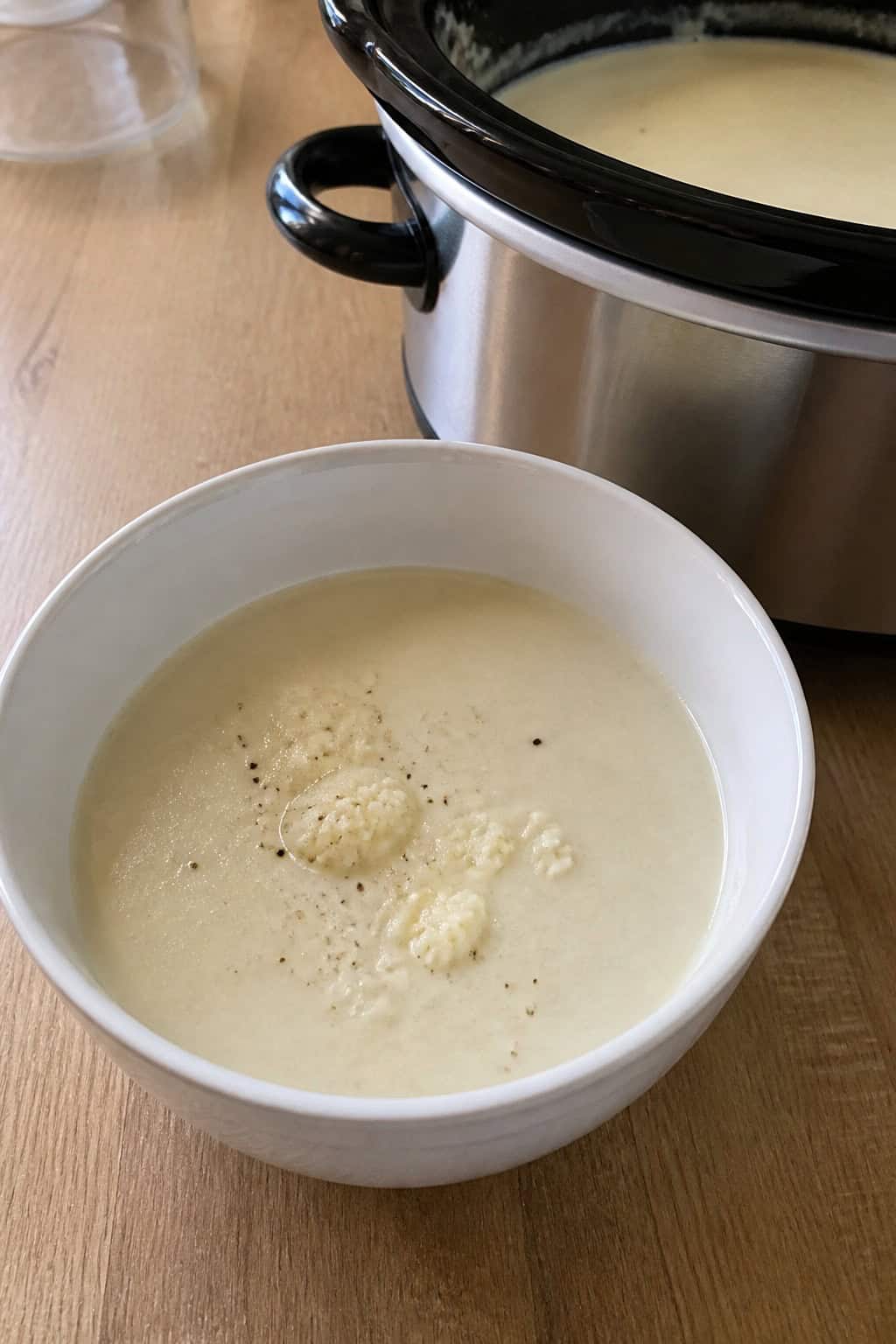A bowl of creamy cauliflower soup garnished with herbs on a wooden table with fresh cauliflower and a slow cooker in the background.