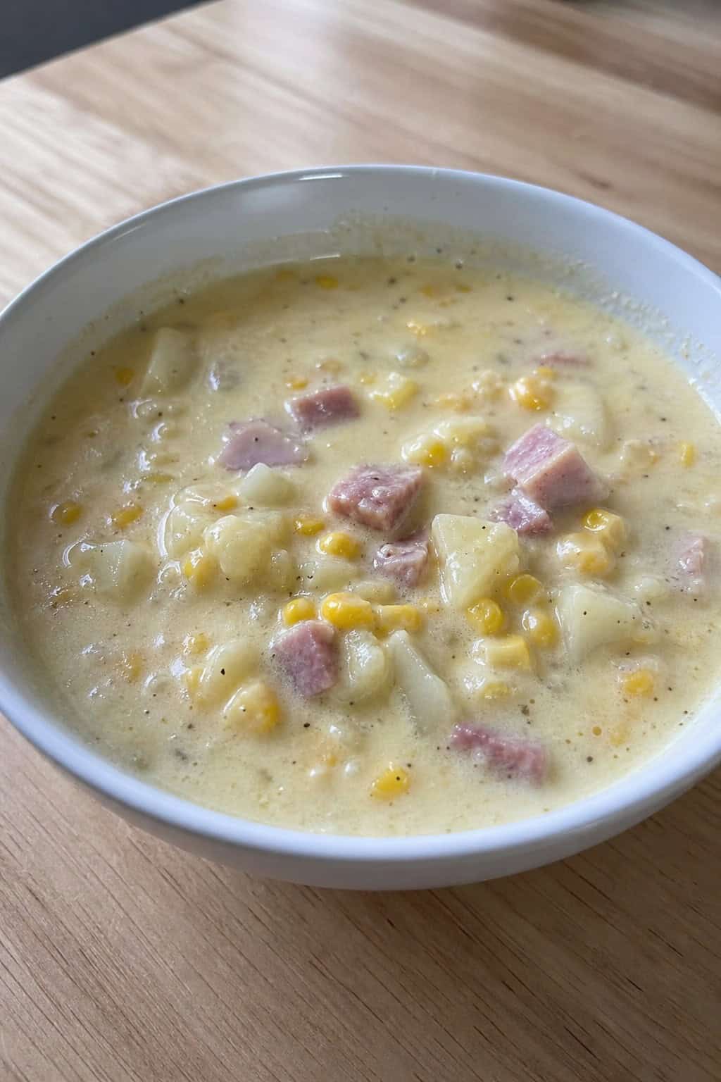 A bowl of creamy corn and ham chowder on a wooden table with fresh corn, ham, and parsley around it.