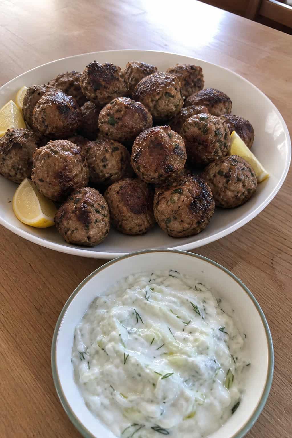 A platter of Greek meatballs with a bowl of tzatziki dip on a table surrounded by fresh herbs and lemon wedges.