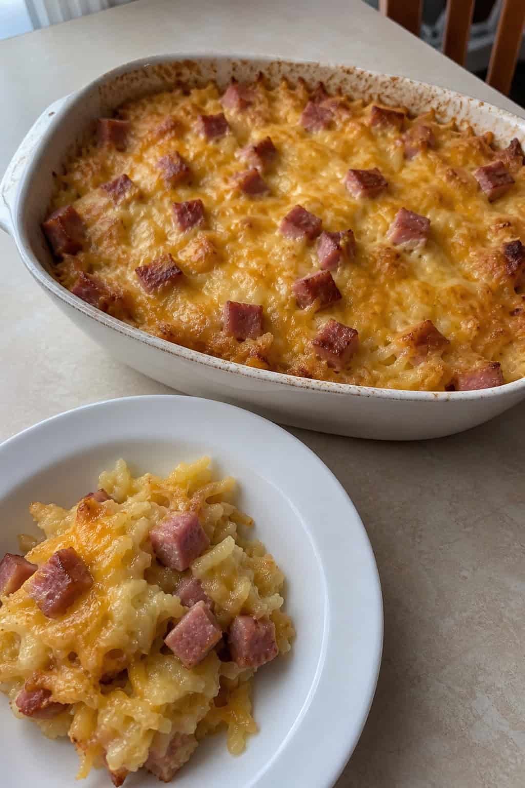 A golden brown ham and hashbrown casserole in a ceramic dish on a wooden table with fresh ingredients nearby.
