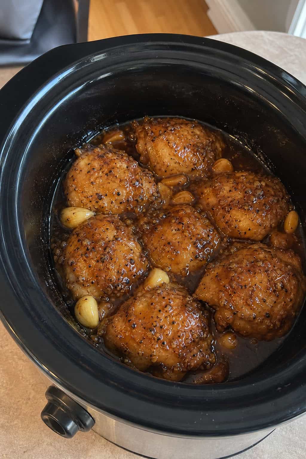 Slow cooker with honey garlic chicken thighs surrounded by fresh garlic, herbs, and honey on a wooden countertop.