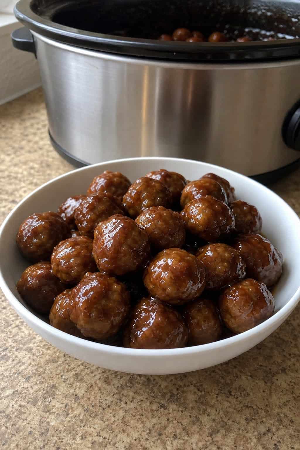 A bowl of honey garlic meatballs on a wooden table with fresh herbs, garlic, and a slow cooker in the background.