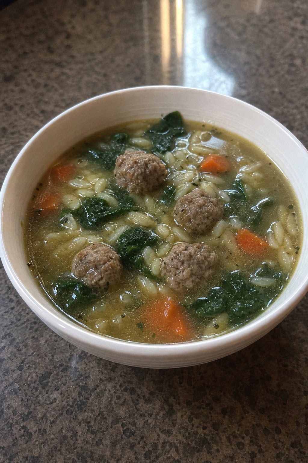 A bowl of Italian Wedding Soup with meatballs, spinach, pasta, and carrots on a wooden table with fresh herbs and bread nearby.