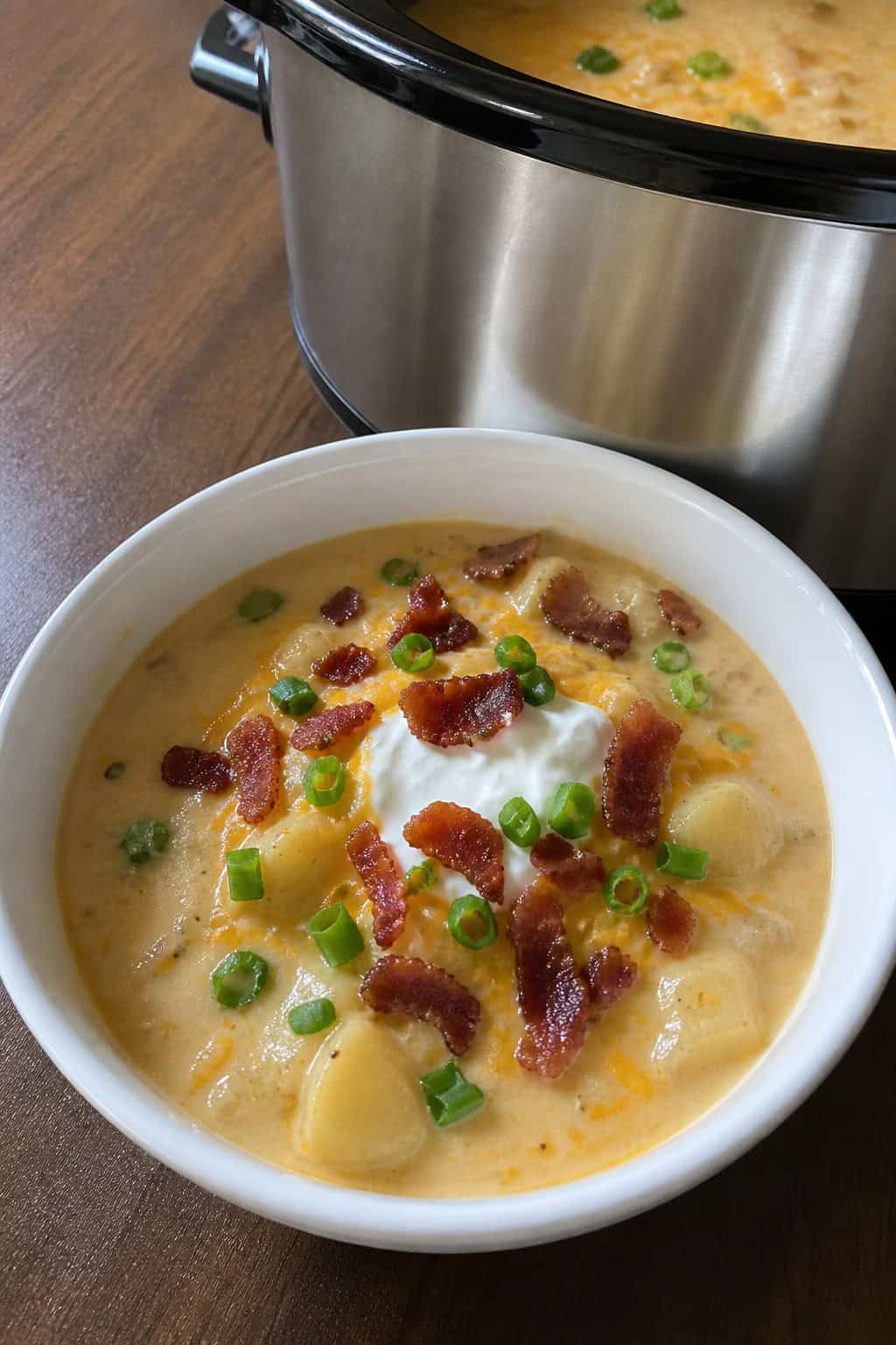 A bowl of creamy loaded baked potato soup topped with cheese, bacon, green onions, and sour cream on a wooden table with fresh ingredients and a slow cooker in the background.