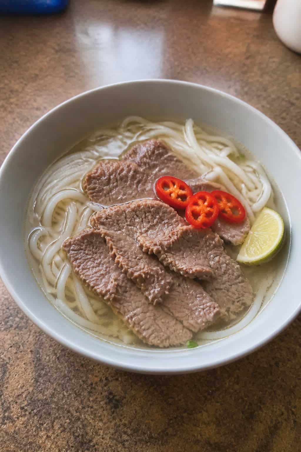 A bowl of steaming pho-style beef noodle soup with fresh herbs and lime on a wooden table.