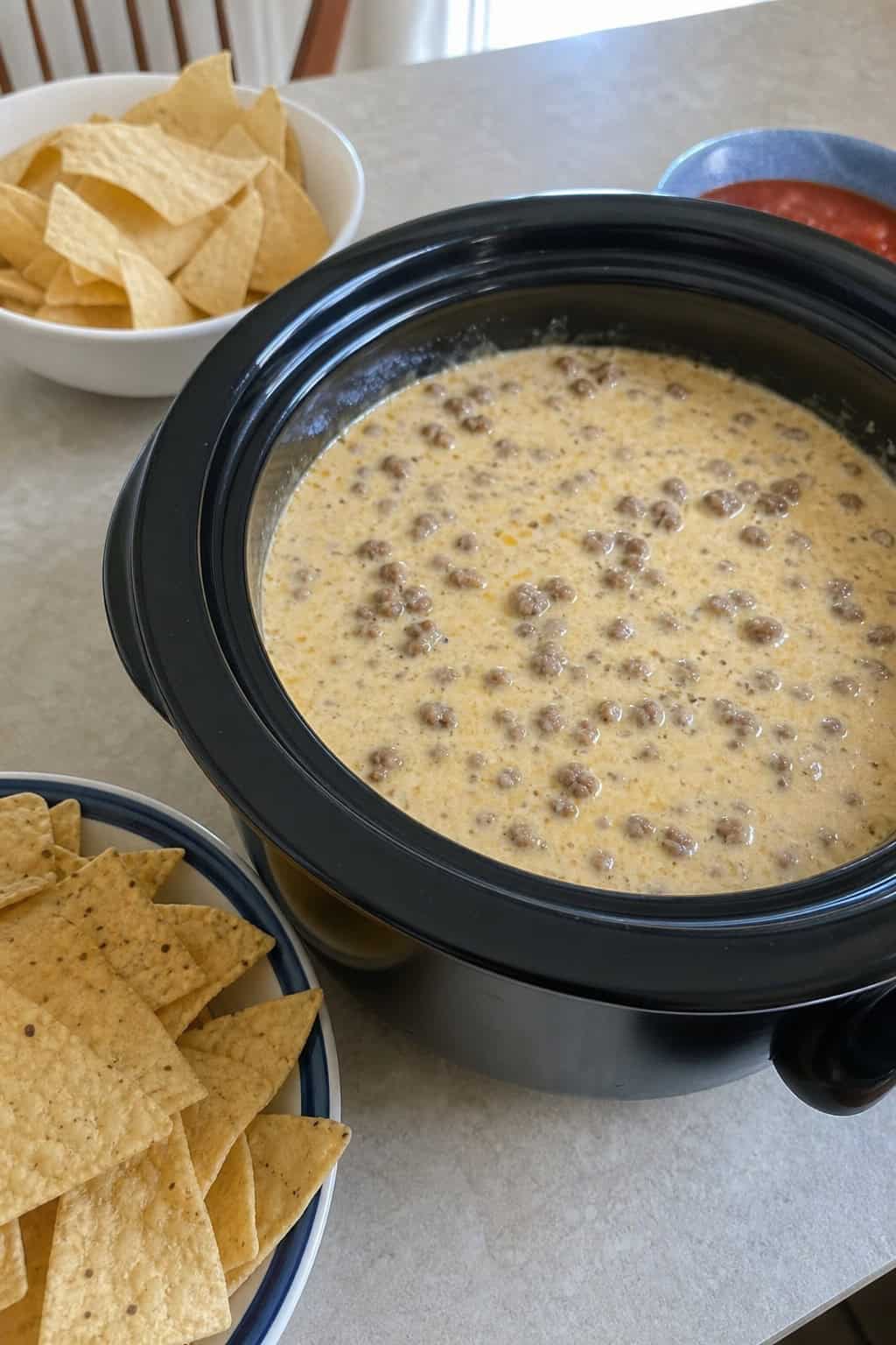 Slow cooker filled with creamy queso blanco and chorizo dip surrounded by bowls of tortilla chips and fresh toppings on a wooden table.