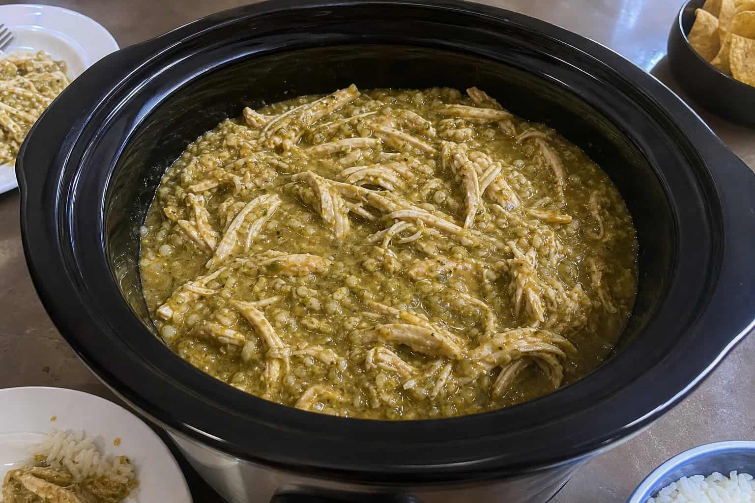 Slow cooker filled with green salsa chicken surrounded by fresh tomatillos, garlic, lime, cilantro, and jalapeños on a wooden countertop.