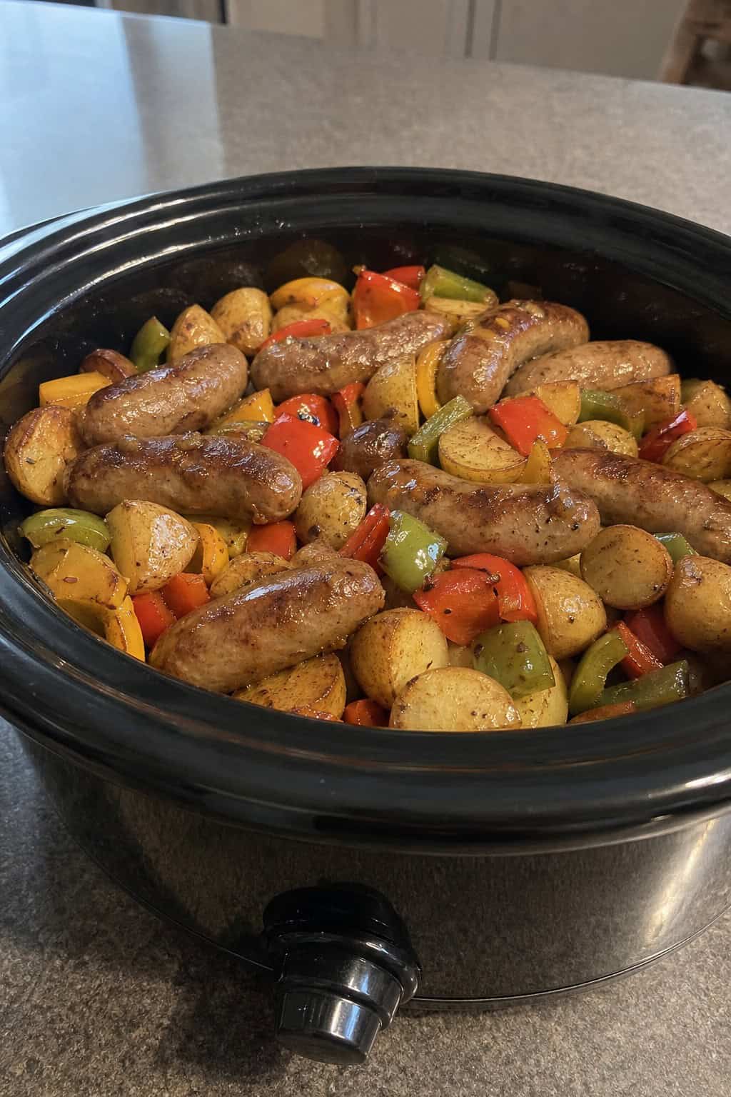 Slow-cooker filled with cooked sausages, bell peppers, and roasted potatoes on a wooden kitchen table with fresh ingredients around it.