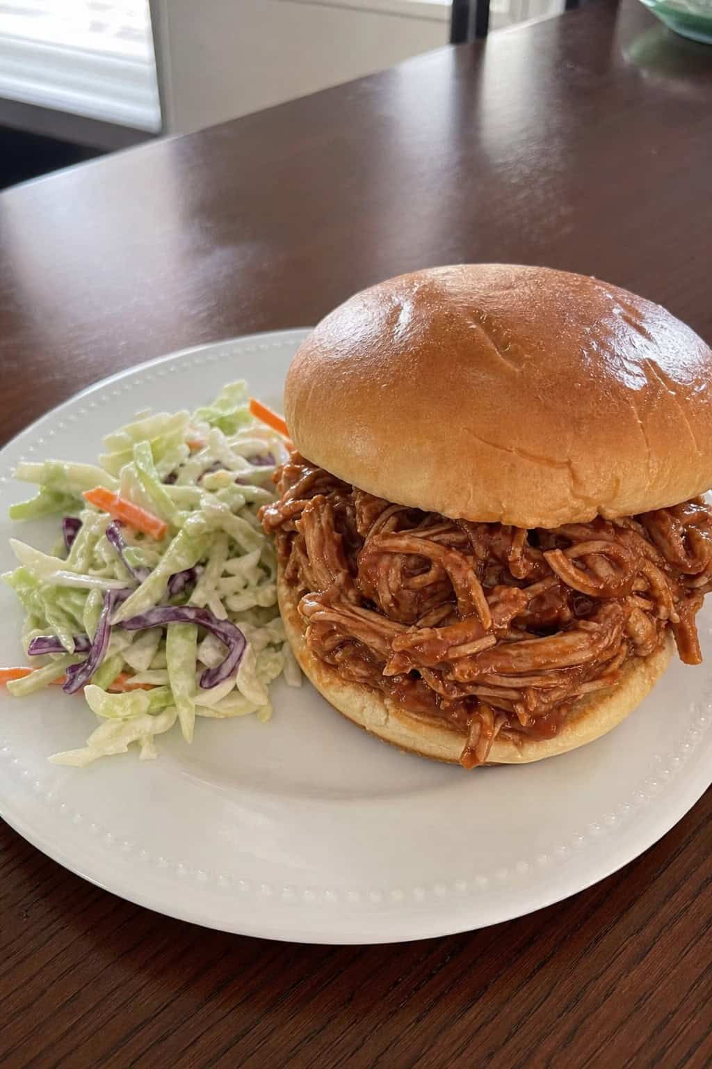 A shredded BBQ chicken sandwich with coleslaw on a wooden table in a bright kitchen setting.