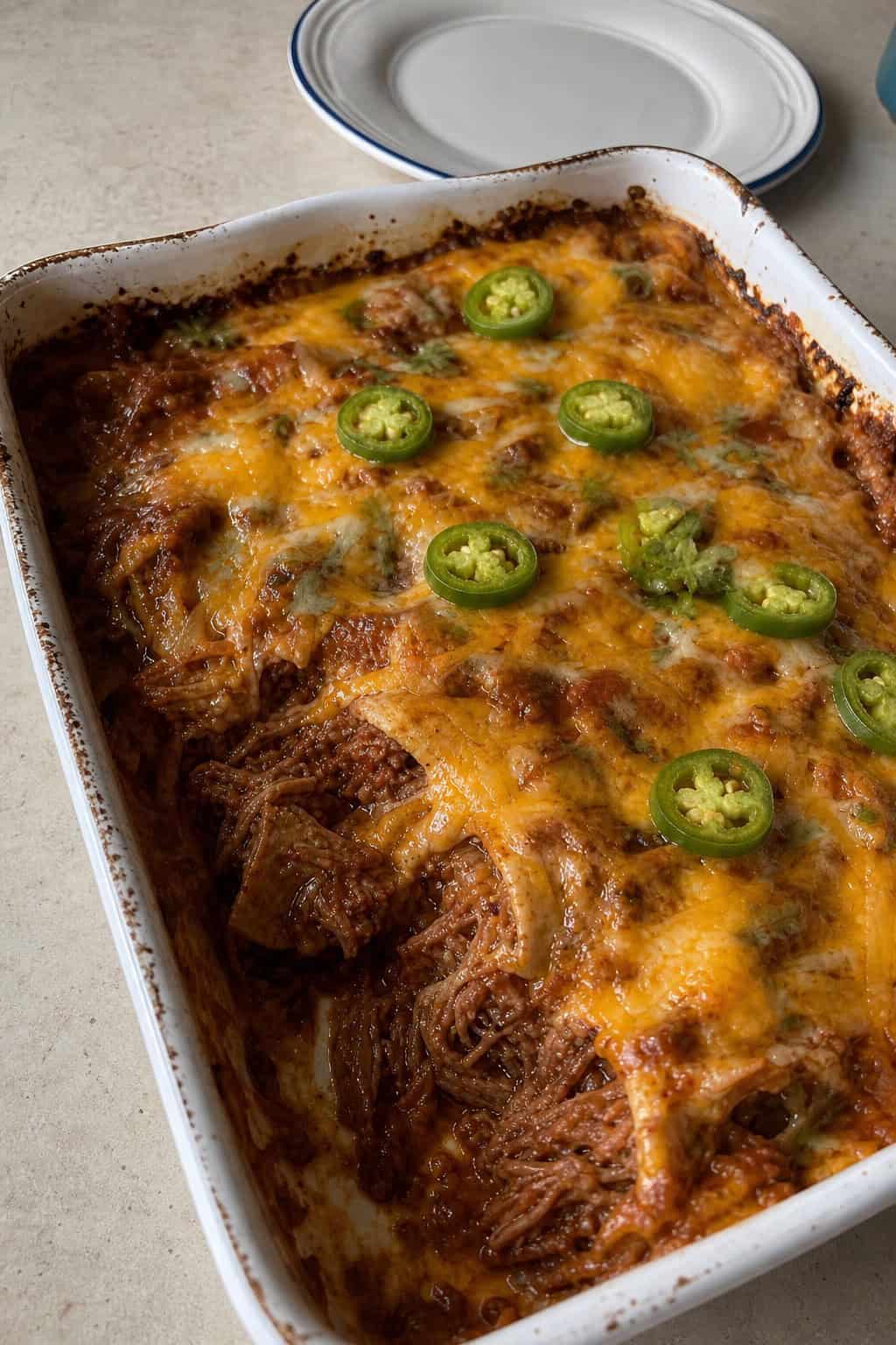 A slow-cooked shredded beef enchilada casserole in a deep ceramic baking dish, with tender pulled beef visible through melted cheese, garnished with fresh cilantro and green jalapeño slices.