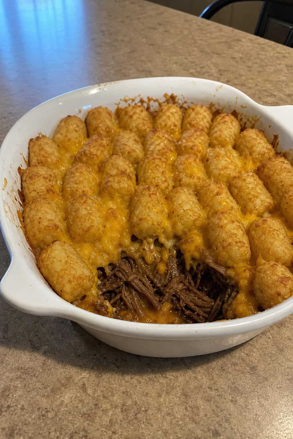 A casserole dish filled with slow cooker shredded beef tater tot casserole, with dark shredded beef and golden tots visible, set on a wooden table with fresh herbs nearby.