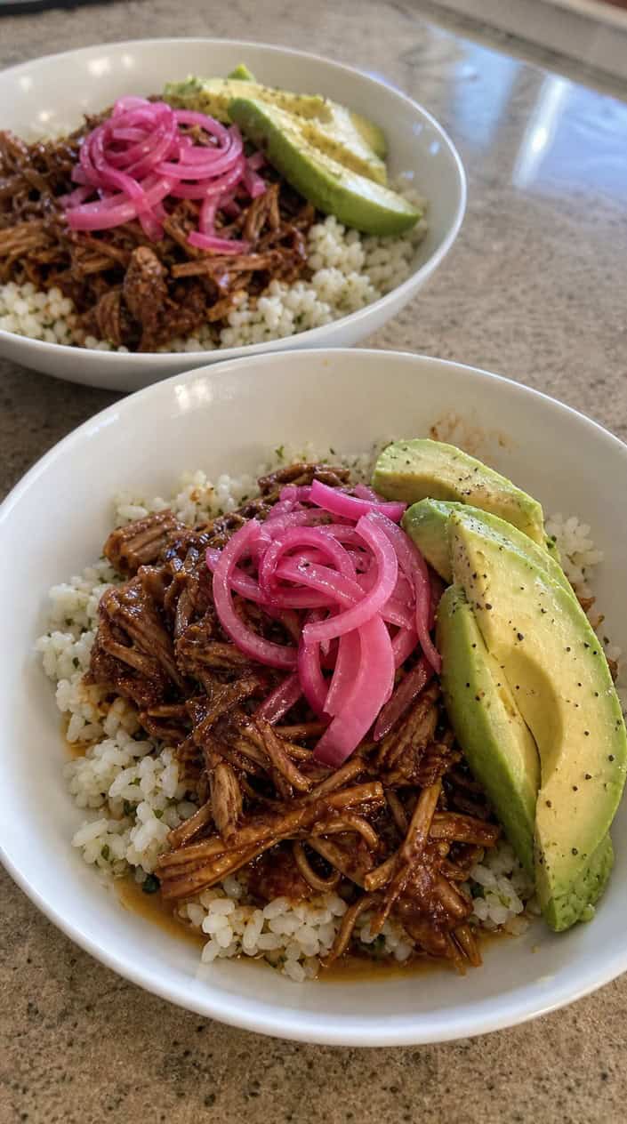 A bowl of smoky adobo-sauced shredded chicken over white rice, layered with bright pink pickled onions, fanned avocado slices, and fresh cilantro, with lime wedges and chopped onion on the table beside it.