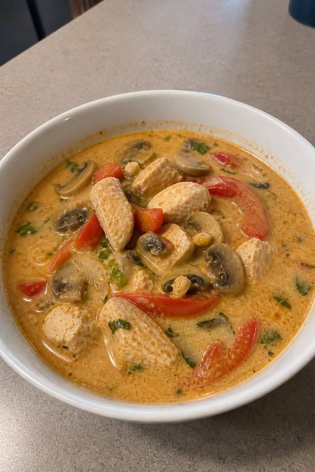 A bowl of Thai coconut chicken soup with chicken, mushrooms, red peppers, and cilantro on a wooden table with fresh ingredients around it.