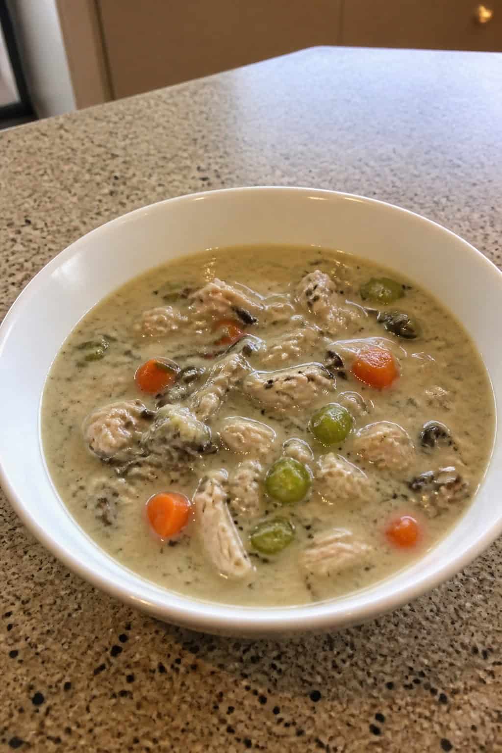 A bowl of turkey and wild rice soup on a wooden table with fresh vegetables and herbs nearby.