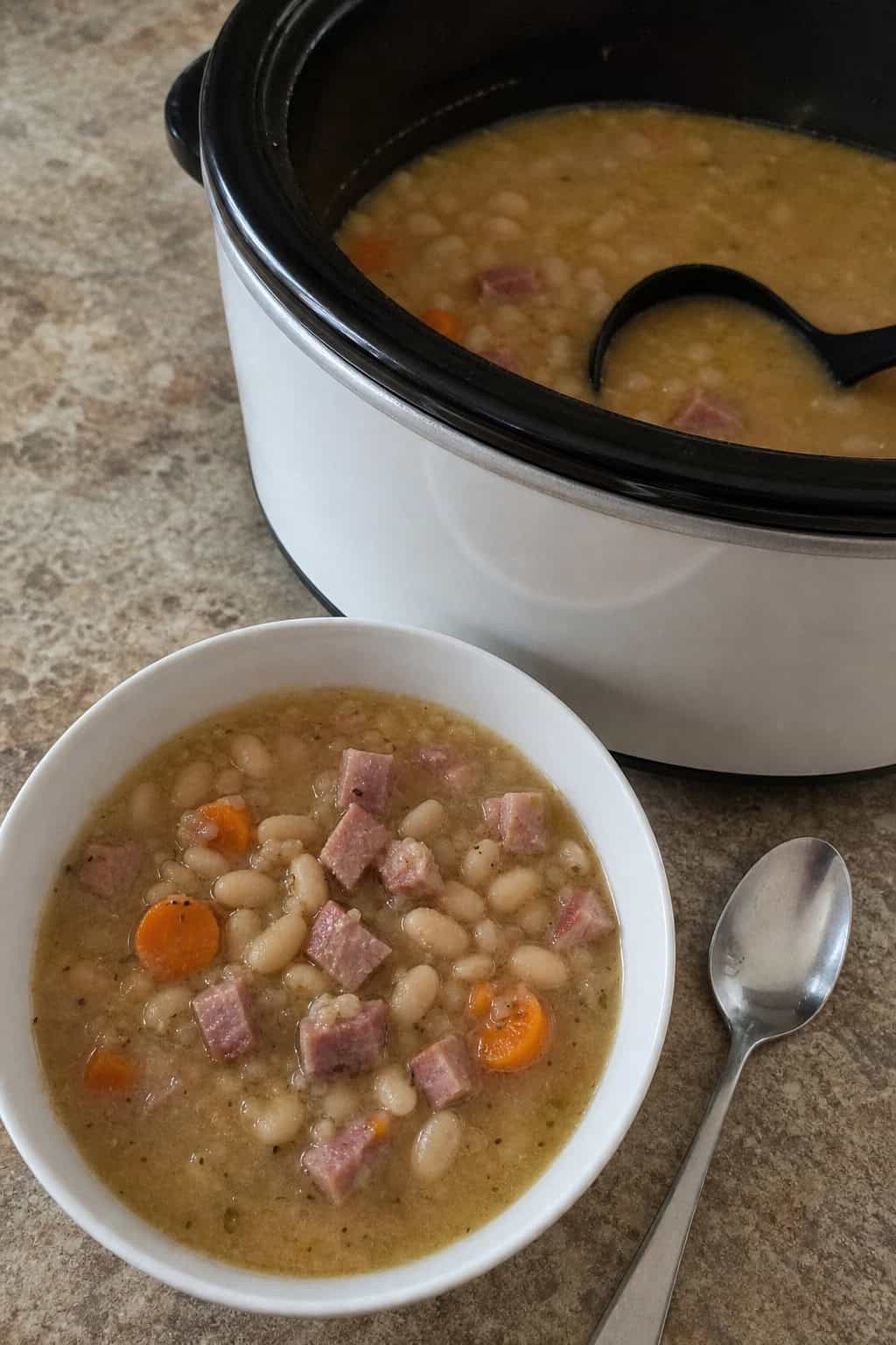 A slow cooker filled with white bean and ham soup on a kitchen countertop surrounded by fresh ingredients like ham, beans, and herbs.