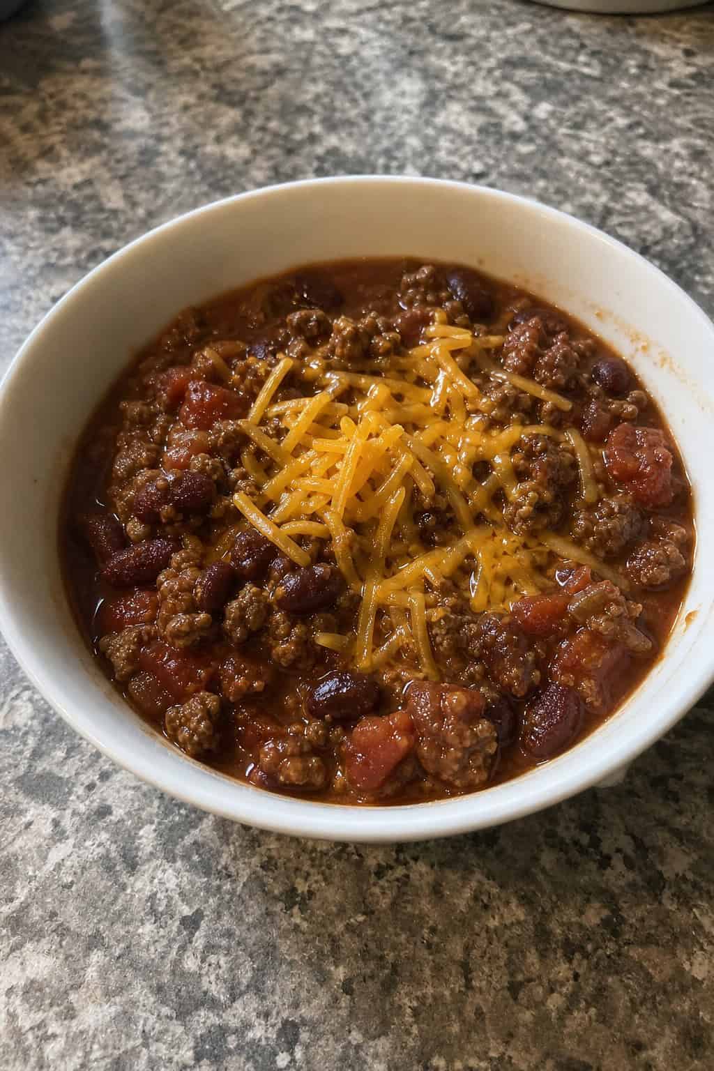 18 Easy Ground Beef Recipes for Weeknight Dinners 13 A bowl of slow-simmered beef chili with beans, cheese, and cilantro on a wooden table with sour cream and lime wedges nearby.