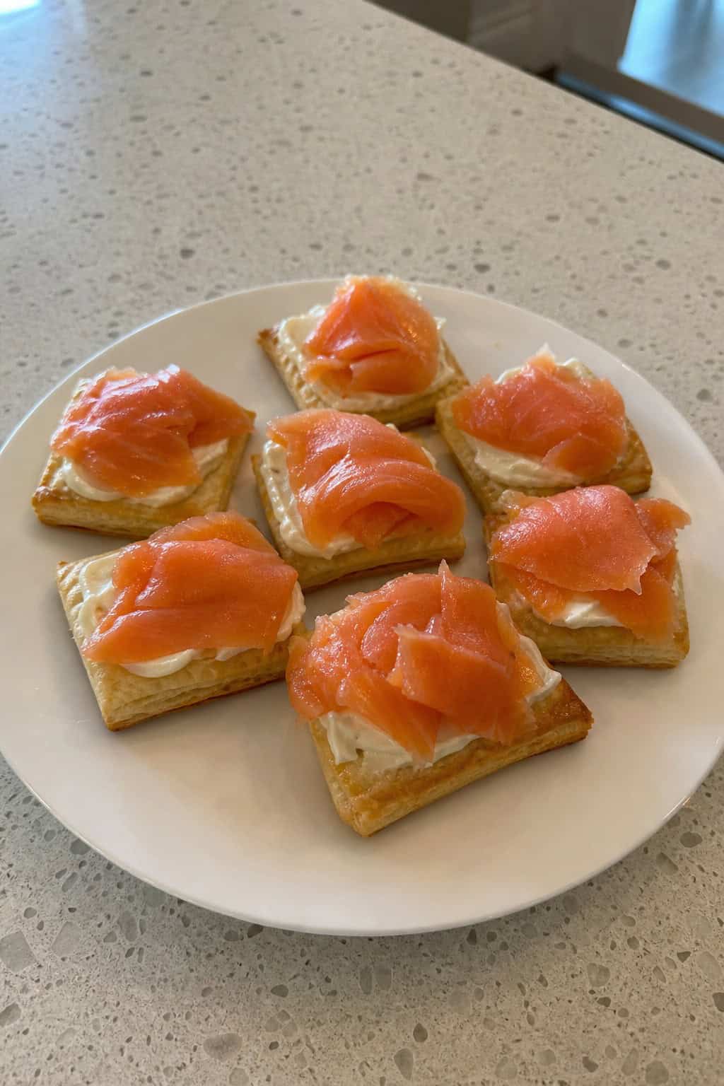 A platter of smoked-salmon cream cheese puff pastry squares garnished with herbs on a wooden board with lemon wedges and a bowl of dip.