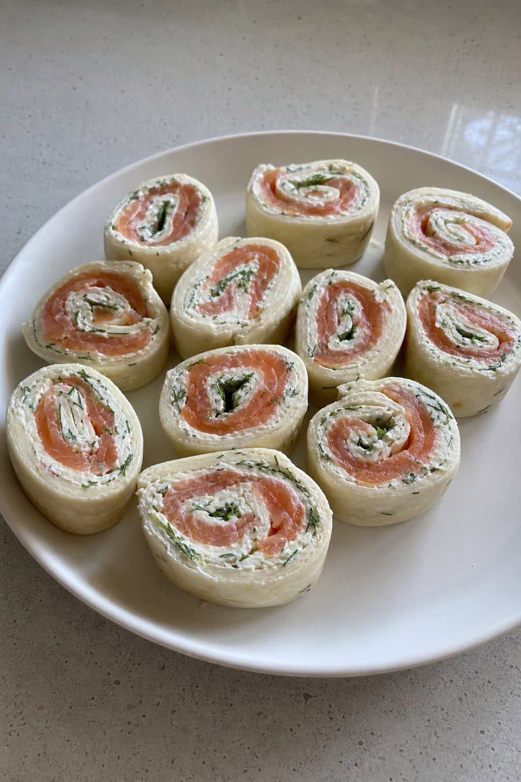 Close-up of smoked salmon dill cream cheese pinwheels on a wooden board showing pale pink salmon slices and thin cucumber rounds inside rolled tortillas, with fresh dill sprigs and lemon wedges alongside.