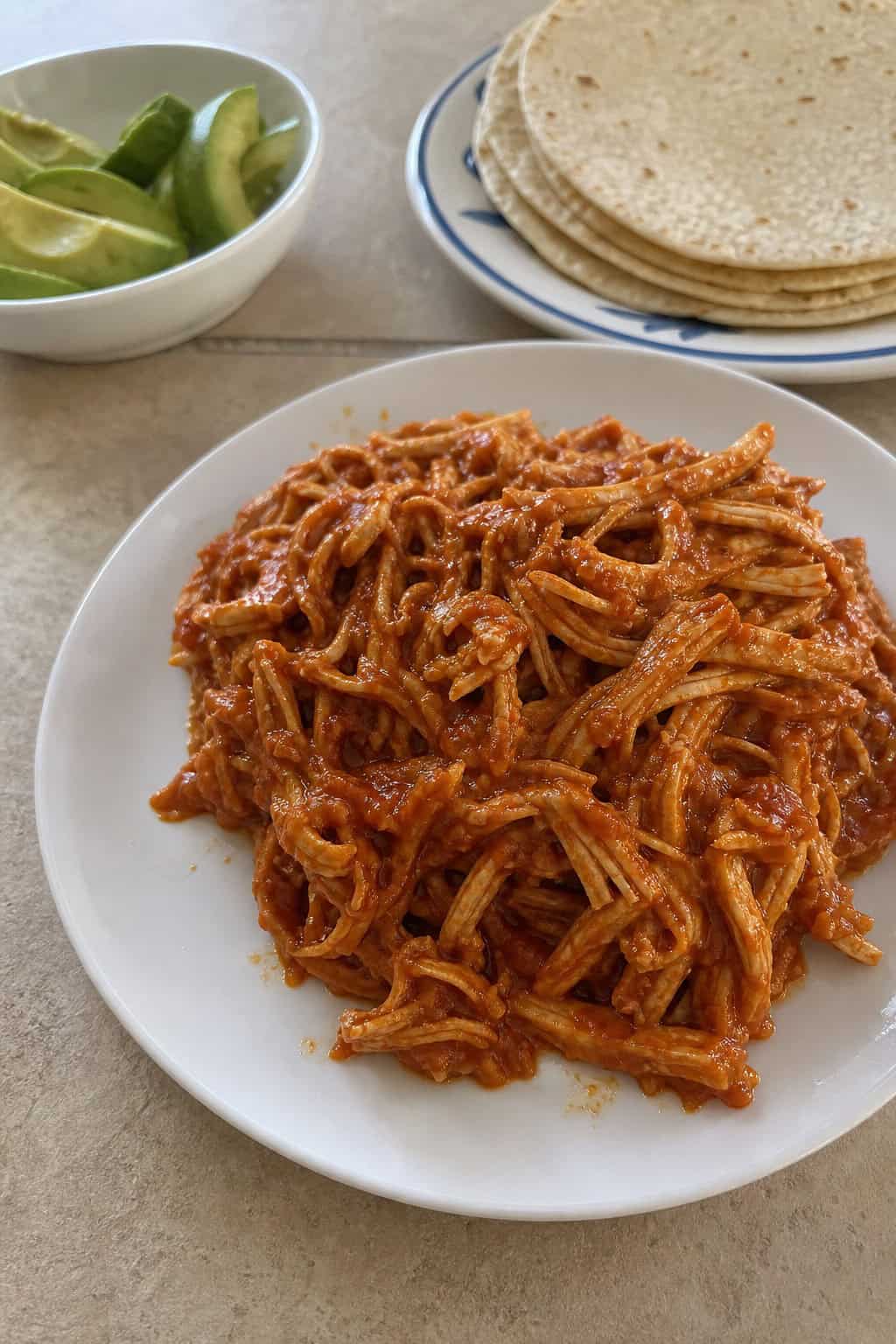 A plate of smoky shredded chicken in red sauce garnished with cilantro, served with avocado slices, lime wedges, onions, and corn tortillas on a wooden table.