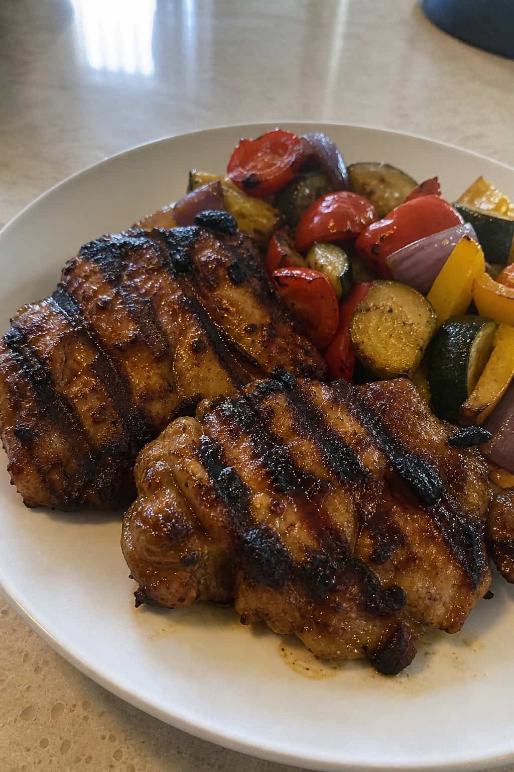 A plate of glazed smoky chipotle honey chicken thighs garnished with herbs and red chili slices, served with roasted vegetables.