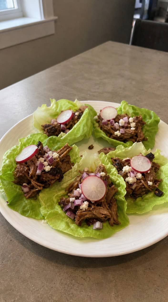 A plate of smoky pork carnitas served in fresh lettuce cups with garnishes of red onion, cilantro, radish slices, and lime wedges.