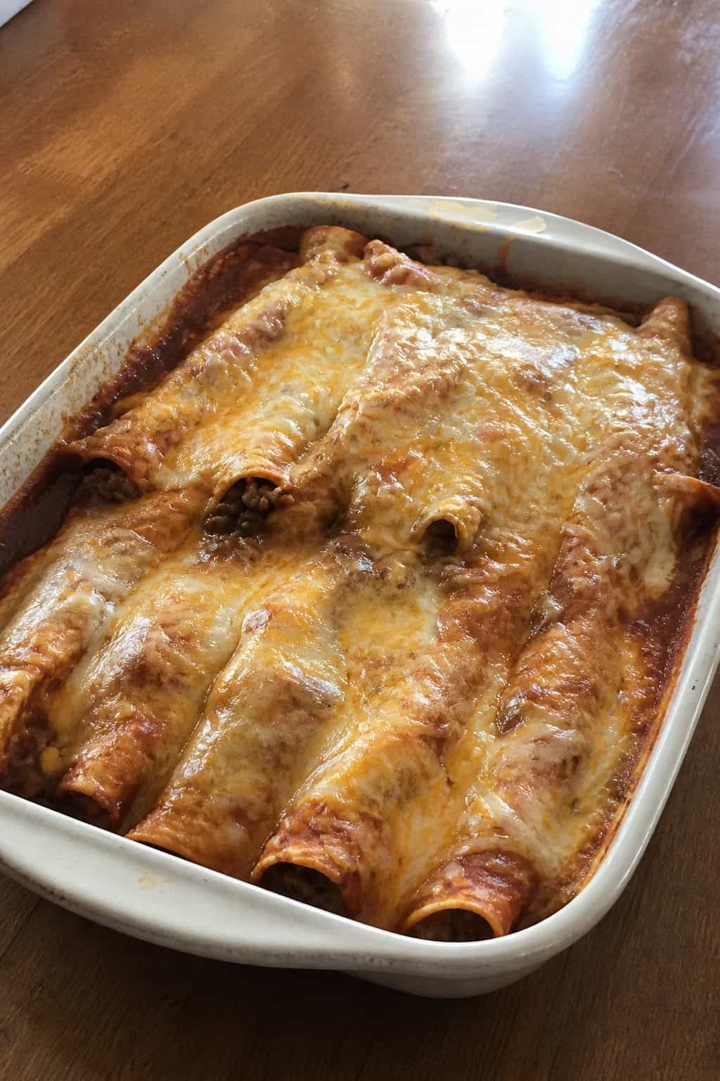 A baked dish of ground beef enchiladas blanketed in bubbling red sauce and melted cheese, garnished with cilantro and sliced green onions, with small bowls of sour cream, tomatoes, and jalapeños visible on the wooden table.
