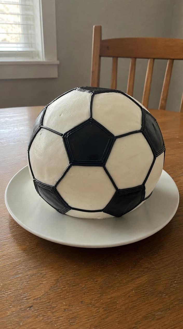 A round birthday cake decorated to look like a soccer ball with black hexagons piped on white icing.