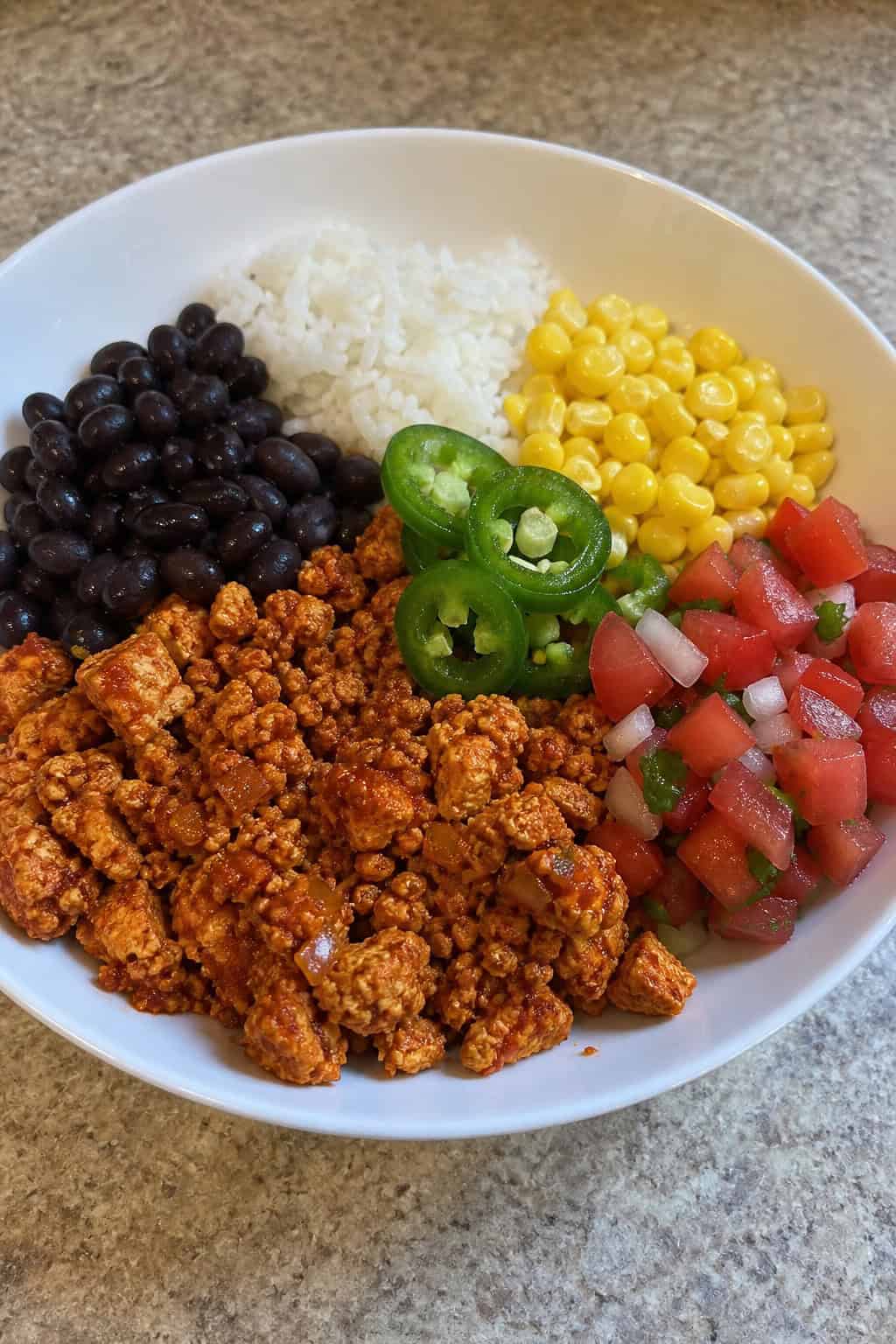 A bowl of seasoned crumbled tofu with rice, beans, corn, and fresh vegetables on a wooden table.