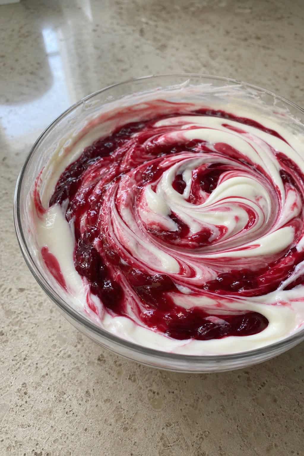A bowl of creamy cheesecake filling swirled with sour cherry sauce, surrounded by fresh sour cherries on a wooden surface.