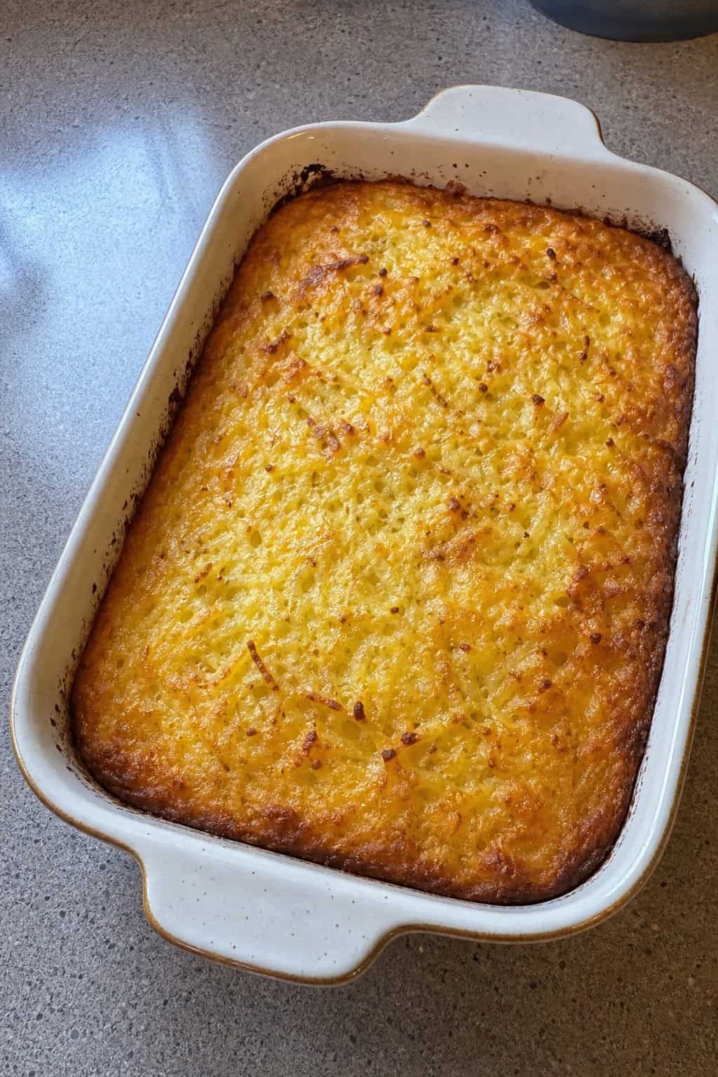 A golden brown sour cream and chive hashbrown casserole in a ceramic dish on a wooden table with fresh chives and a serving spatula nearby.
