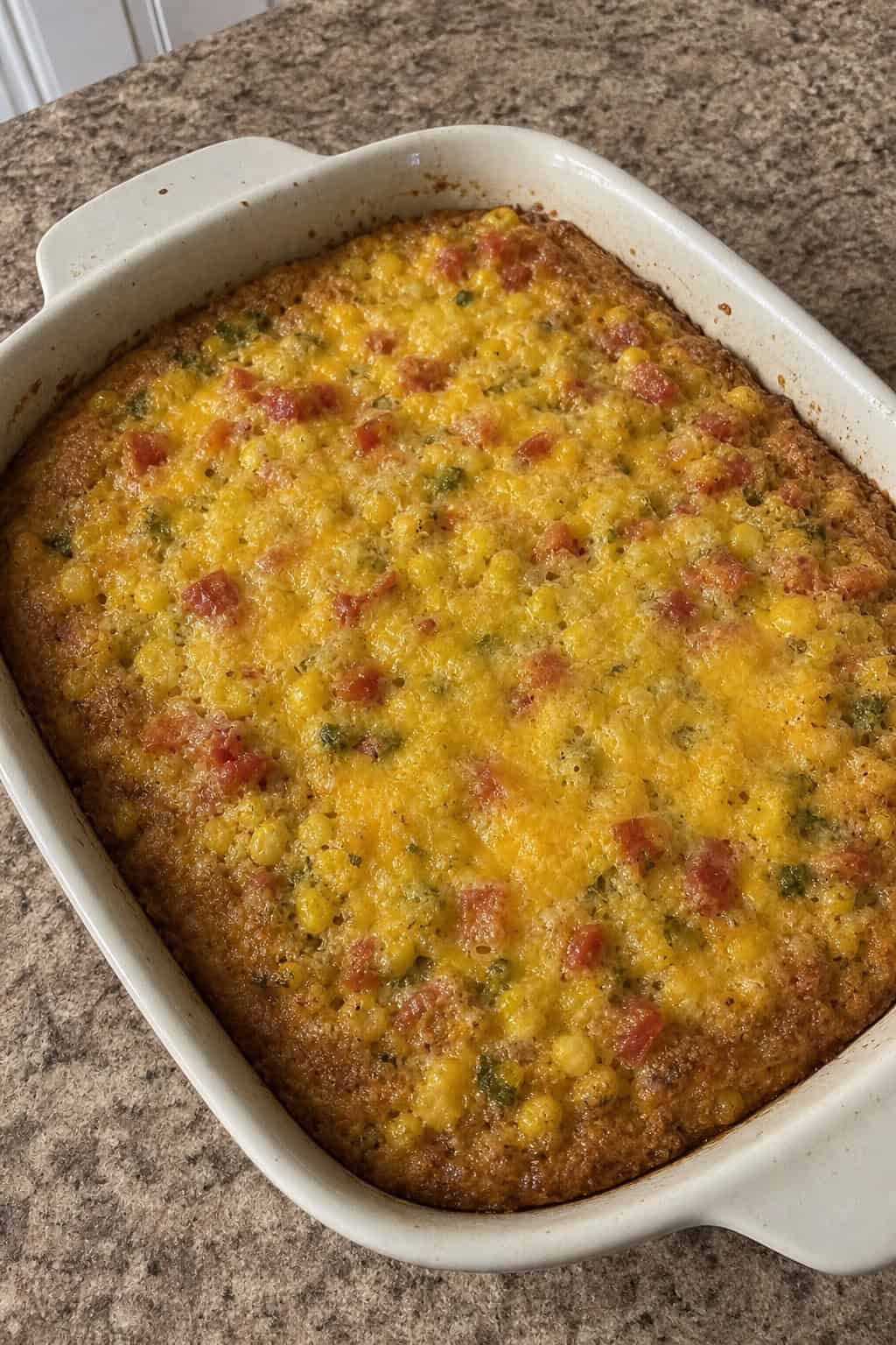 A baked corn casserole flecked with red tomato and bright green cilantro in a white ceramic dish on a wooden table, with a can of Rotel and a bunch of fresh cilantro nearby.
