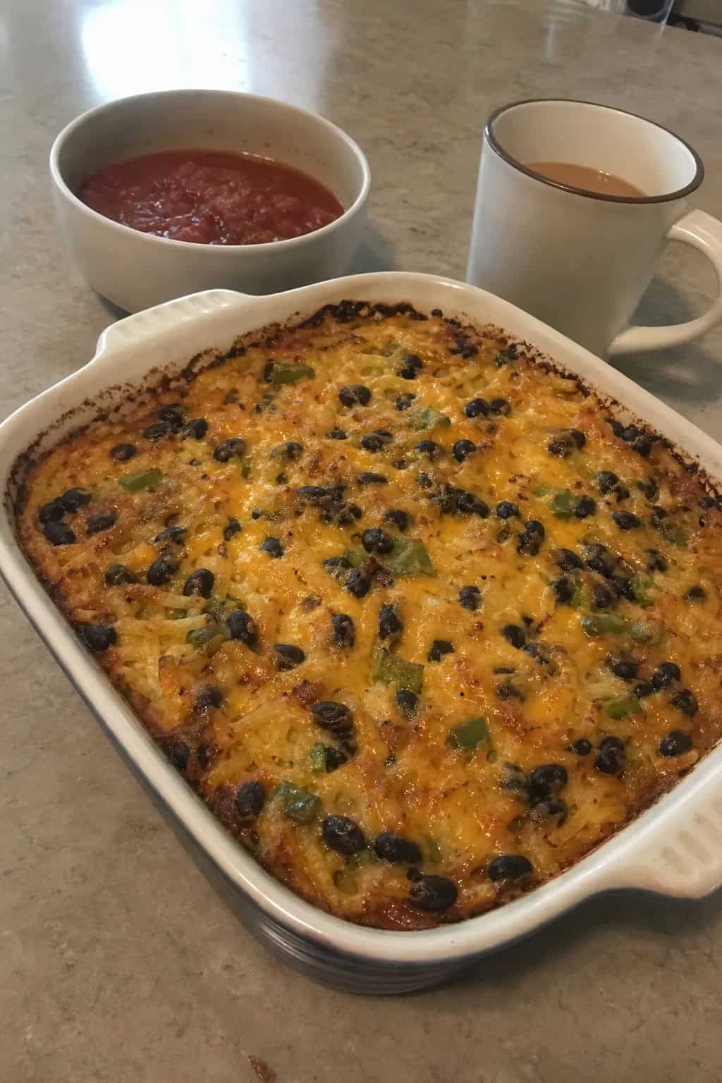 A baked Southwestern hashbrown casserole with visible black beans and a golden cheese top in a white ceramic dish, on a wood table with a fork and folded napkin beside it.