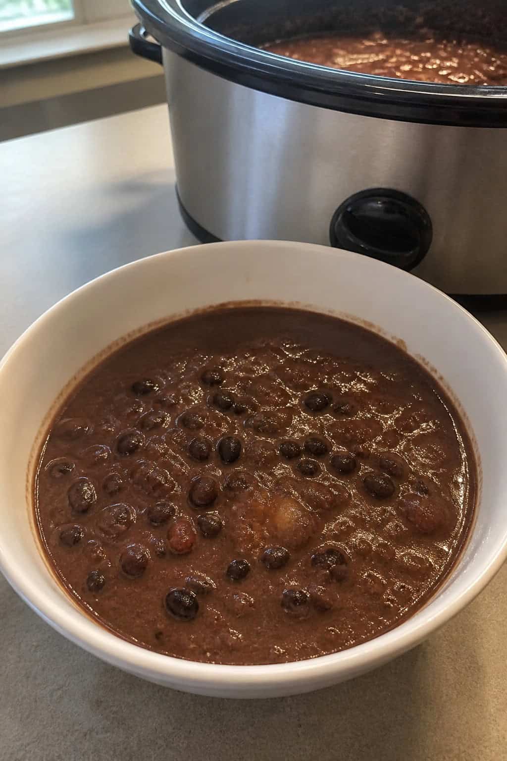 A steaming bowl of black bean and chipotle soup garnished with cilantro and lime on a wooden table with ingredients and a slow cooker in the background.