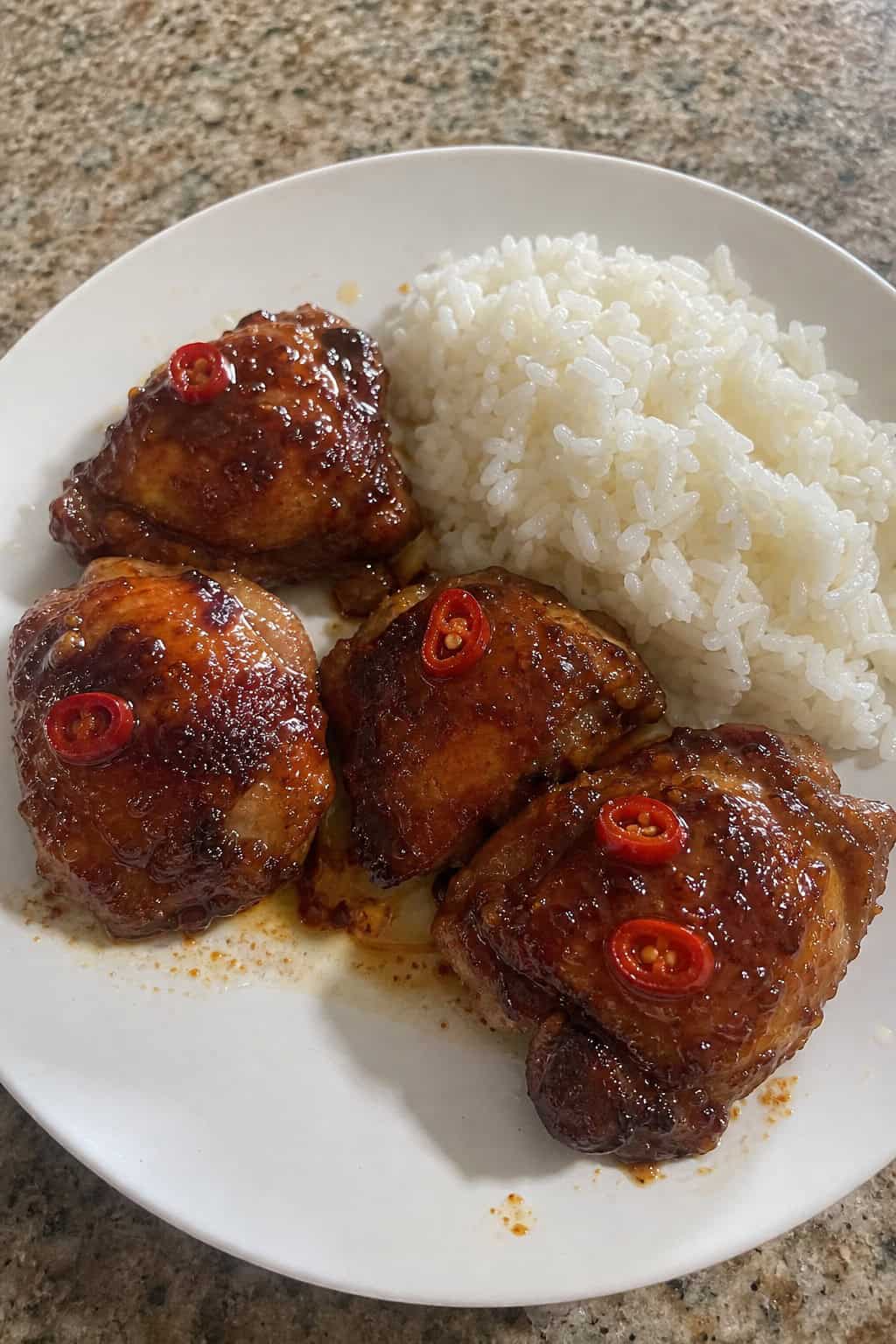 Honey sriracha glazed chicken thighs on a wooden table plated over white rice, with a deeply lacquered reddish-orange sauce, sliced fresh chili peppers, and scattered herb leaves on top.