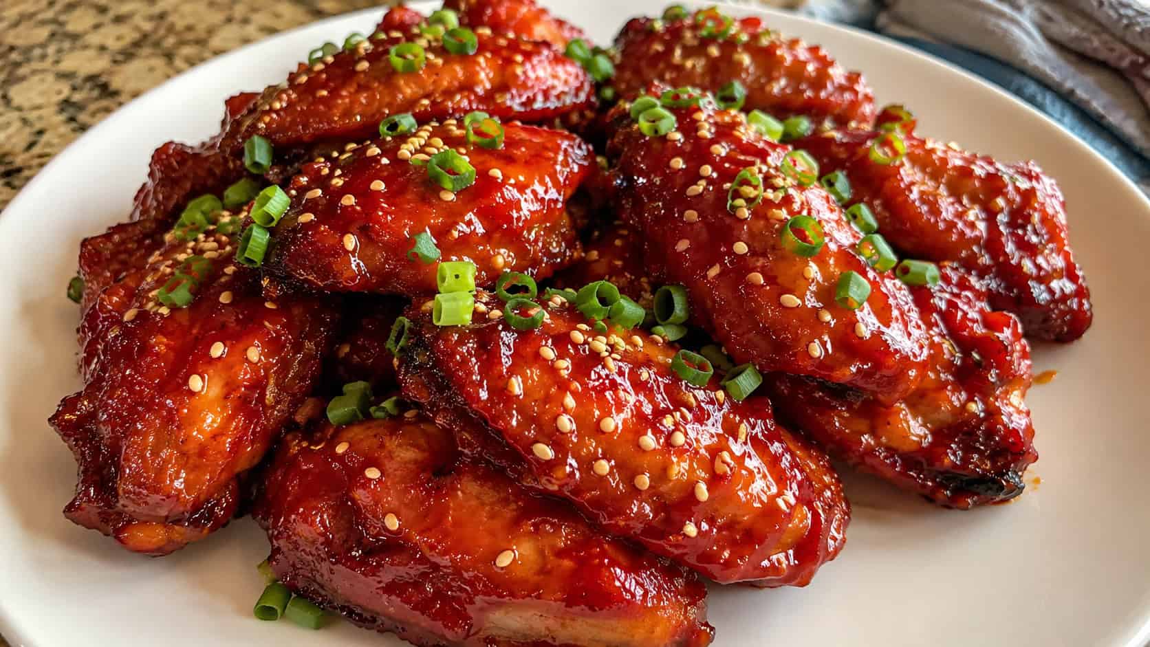 finished Korean-style air fryer chicken wings piled on a plate, glossy deep red sticky gochujang glaze, sliced scallions and toasted sesame seeds scattered on top, warm natural light, casual home cooking