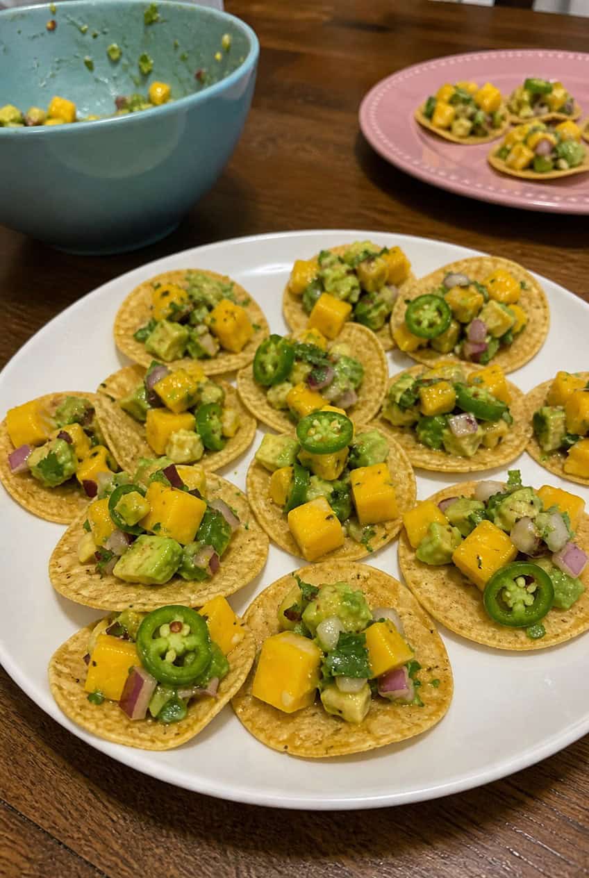A wooden board with sturdy scoop-style tortilla chips each filled with a spoonful of chunky mango and avocado salsa showing bright orange mango, pale green avocado, and flecks of red onion, with lime wedges and fresh cilantro scattered around.