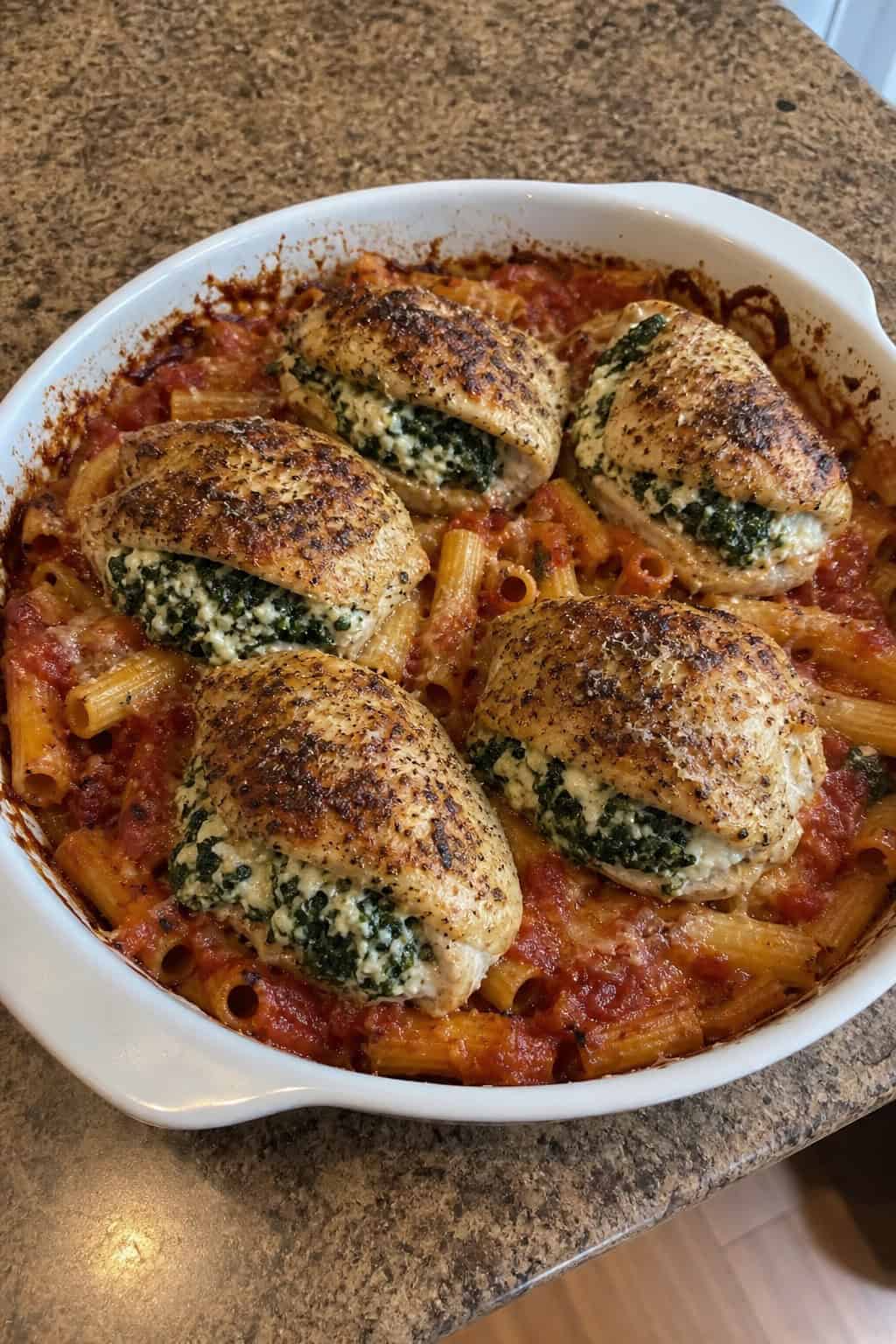 A baked pasta dish with stuffed chicken breasts topped with melted cheese and spinach in a ceramic baking dish on a wooden table.