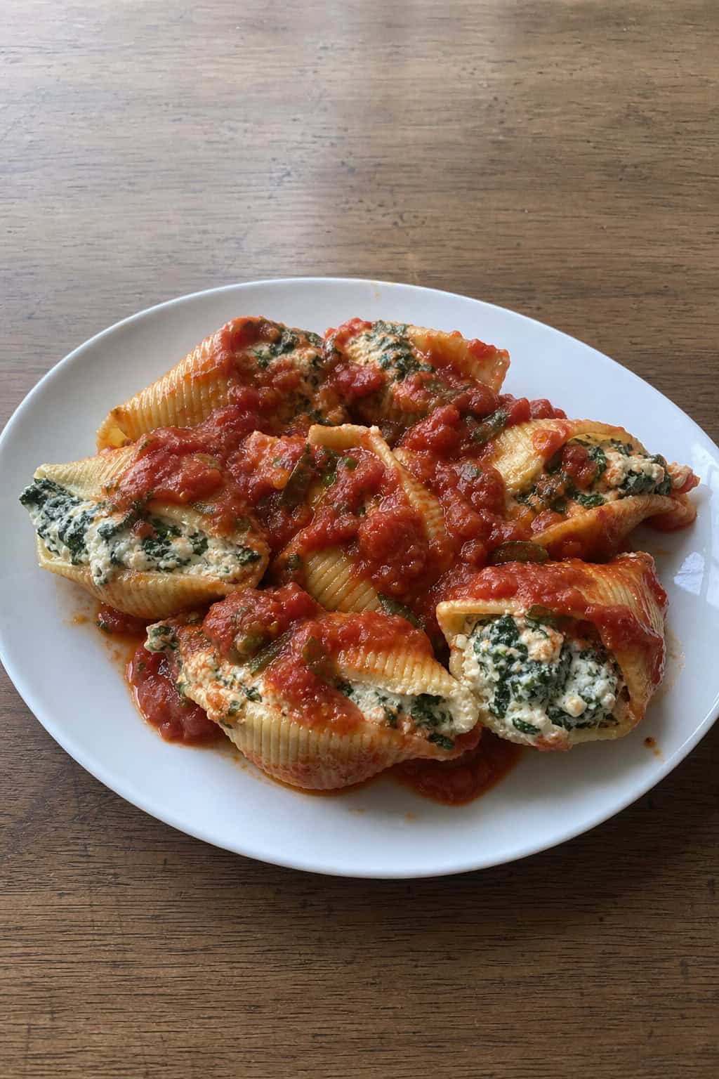 A plate of spinach and ricotta stuffed pasta shells in tomato sauce garnished with basil stems on a wooden table with fresh basil leaves and a glass of white wine nearby.