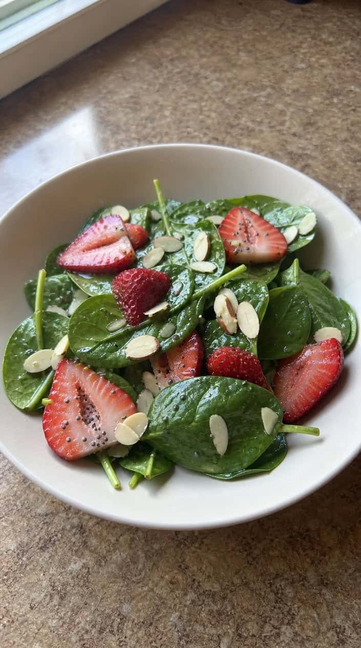 A bowl of spinach and strawberry salad with poppy seed dressing on a wooden table.