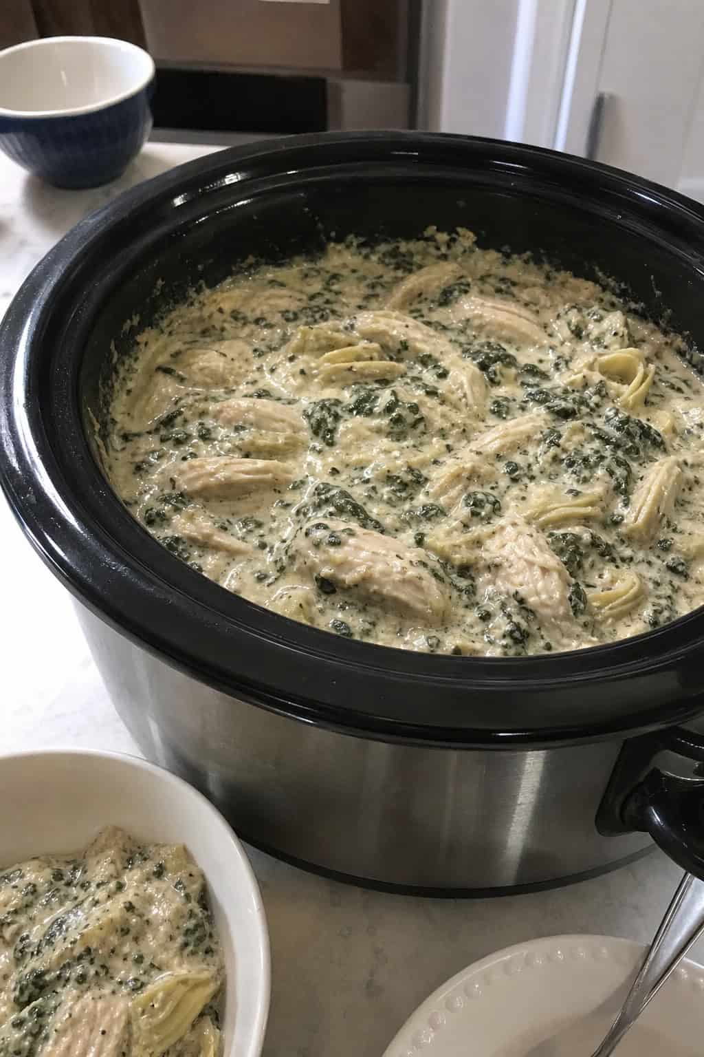 A slow cooker filled with creamy spinach artichoke chicken surrounded by fresh spinach, artichokes, and garlic on a wooden countertop.